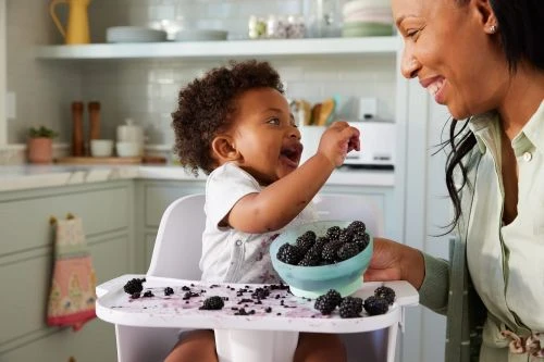 Smiling mother watches her baby in a high chair eating blackberries in a bright kitchen, creating a heartwarming, family moment.