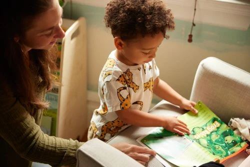 Child reads 'Frog Belly Soap' book with assistance in a cozy room; focus on literary engagement and familial support.