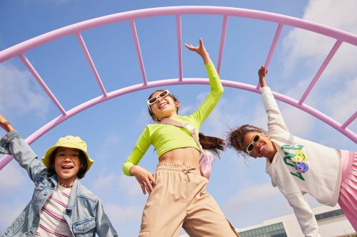 Three children play on a pink climbing frame against a blue sky backdrop, captured from a low angle on a sunny day.
