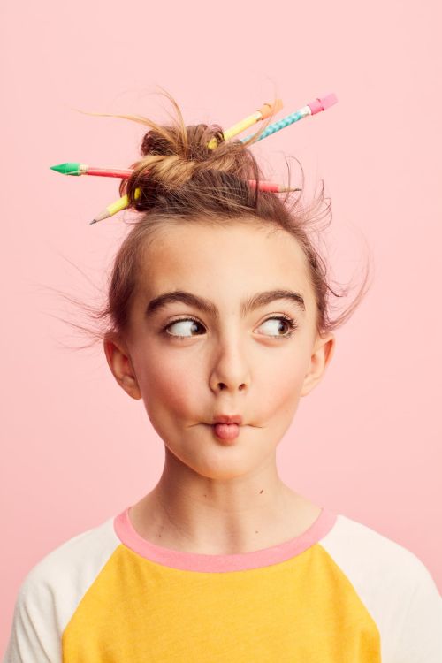 Young girl with pencils in her hair poses against a pink backdrop, wearing a yellow and white shirt. Creative back-to-school fashion shoot.