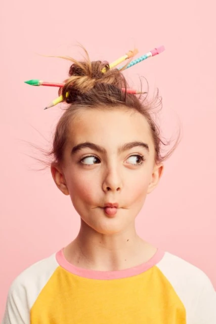 Young girl with pencils in her hair poses against a pink backdrop, wearing a yellow and white shirt. Creative back-to-school fashion shoot.