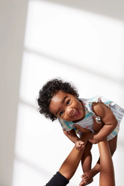 Happy toddler girl in a colorful dress being held in the air by an adult against a white wall with a window casting light patterns.