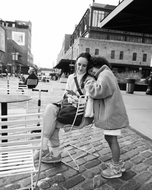 Black and white outdoor shot of two women; one seated looking at a phone, the other standing with her head resting on the other's shoulder.