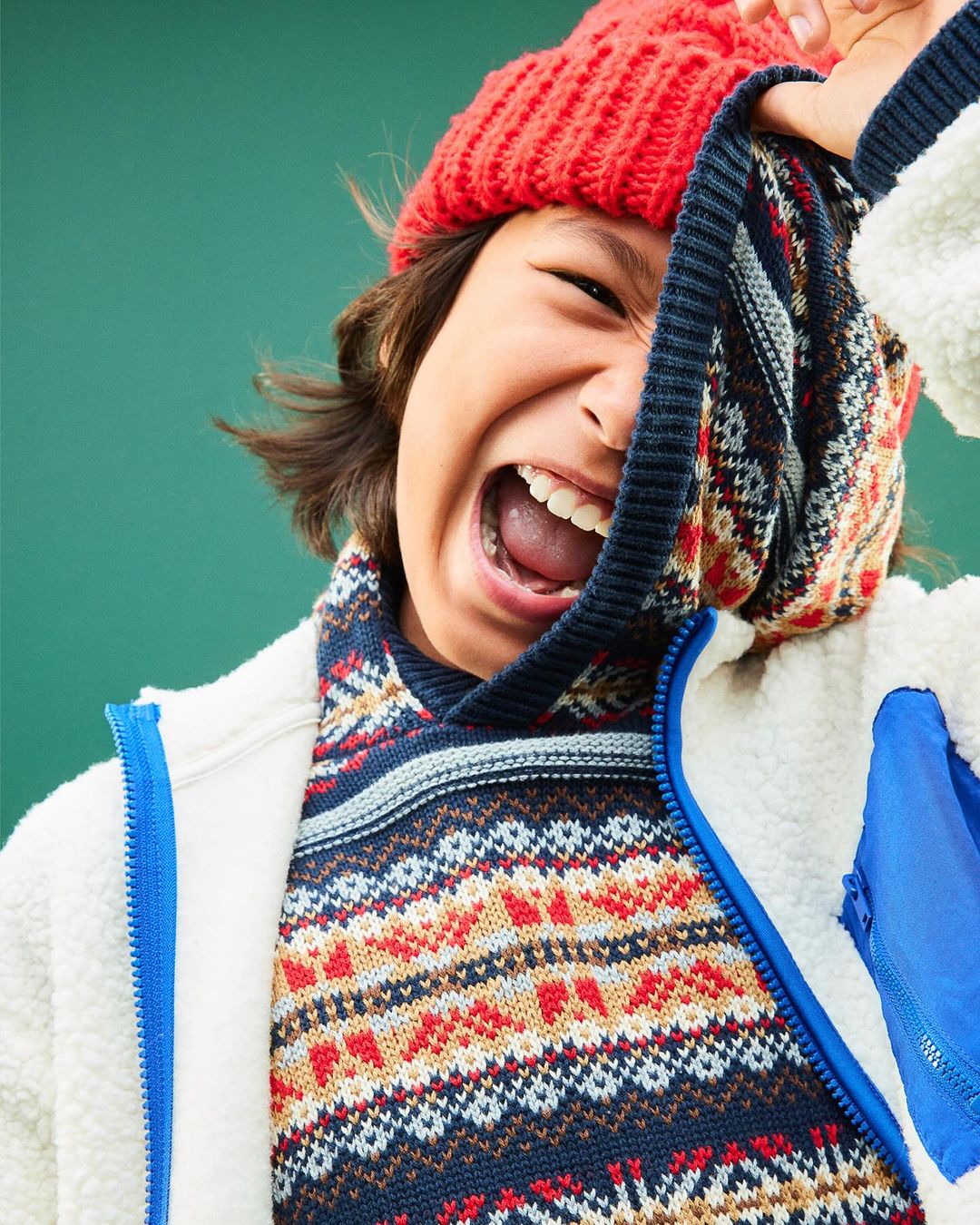 Joyful child with a bright red beanie, pulling a patterned sweater up to cover half the face against a solid green background.