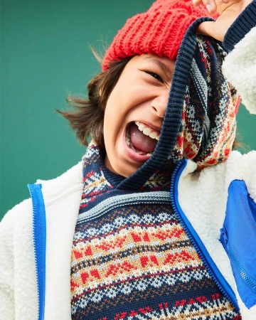 Joyful child with a bright red beanie, pulling a patterned sweater up to cover half the face against a solid green background.