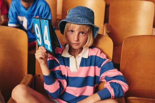Blond child in blue and pink striped shirt and bucket hat holding number card sits in stadium seat; stadium seating background.