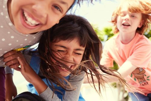 Three cheerful children laughing outdoors in bright sunlight, close-up shot focusing on their joyful expressions and dynamic poses.