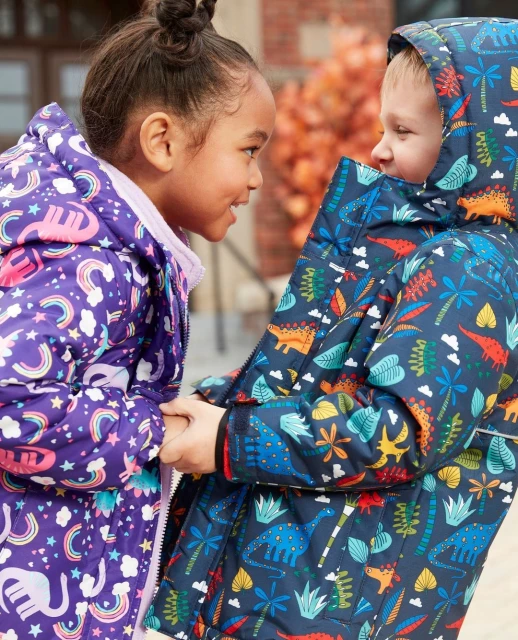Two kids facing each other holding hands, wearing patterned jackets with hoods, standing outdoors against a neutral backdrop on a sunny day.