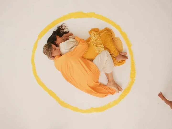Two people lying on a white surface inside a painted yellow circle, creating a cozy and artistic scene with warm tones.