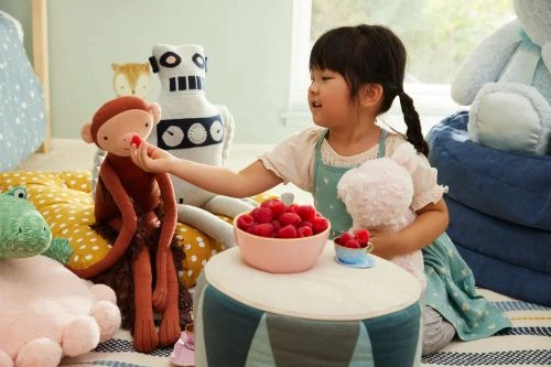 A young girl sitting on a striped rug, holding a teddy bear, and offering a raspberry to a stuffed monkey, with other toys nearby.