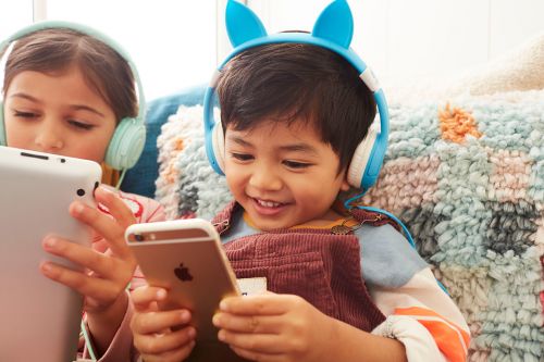 Two children with headphones use a tablet and smartphone, sitting indoors near a patterned throw pillow. They appear happy and engaged.