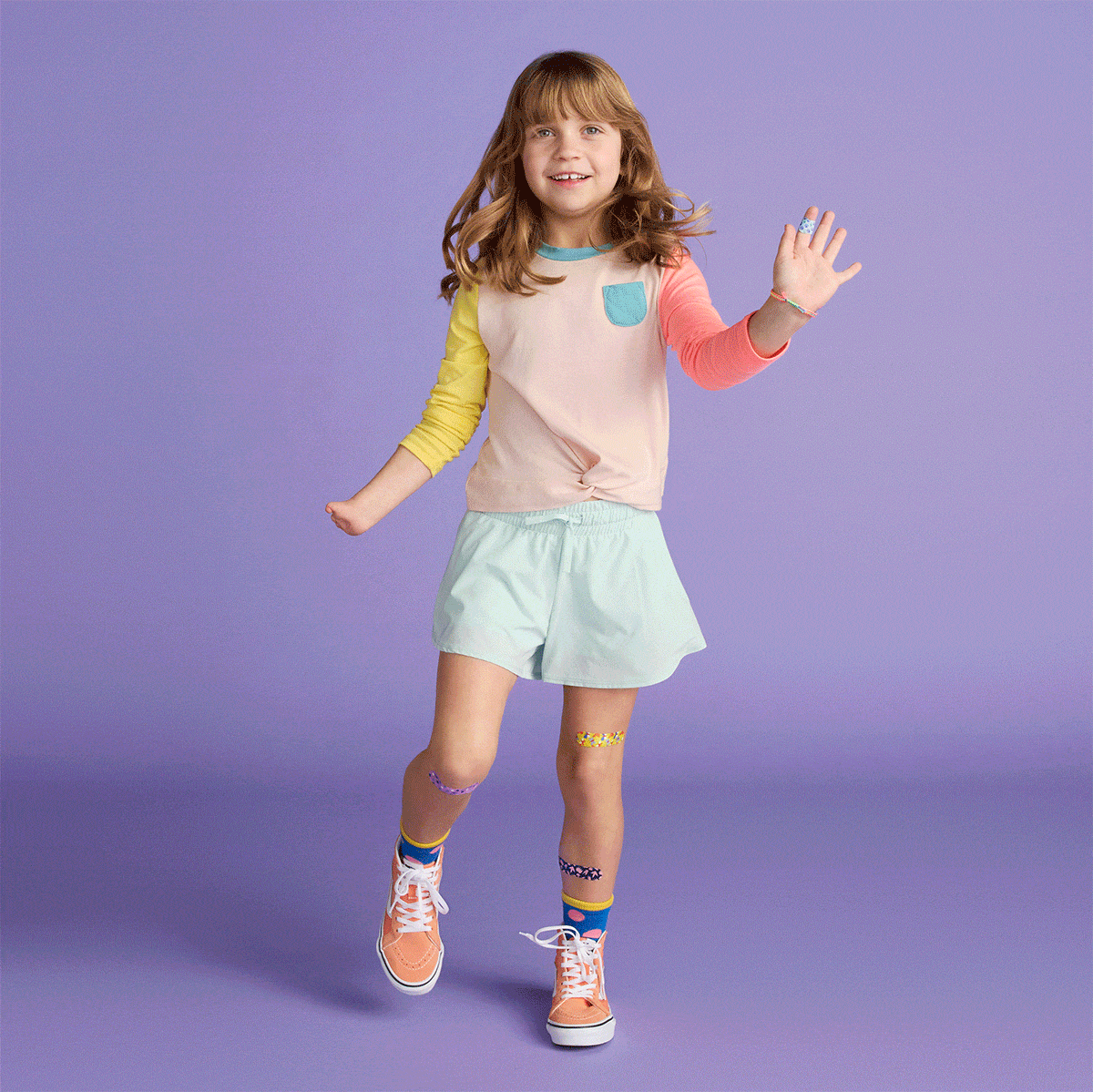 A young girl in colorful clothes smiles and waves against a solid lavender background in a studio shot for children's fashion.