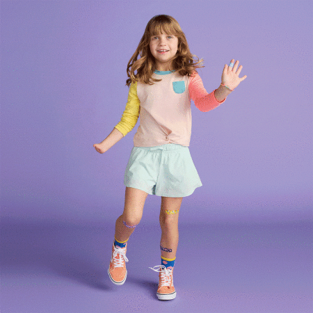 A young girl in colorful clothes smiles and waves against a solid lavender background in a studio shot for children's fashion.