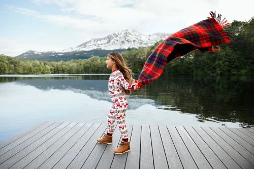 Young girl in pajamas on a wooden dock, holding a red plaid blanket by a lake with snow-capped mountains in the background.