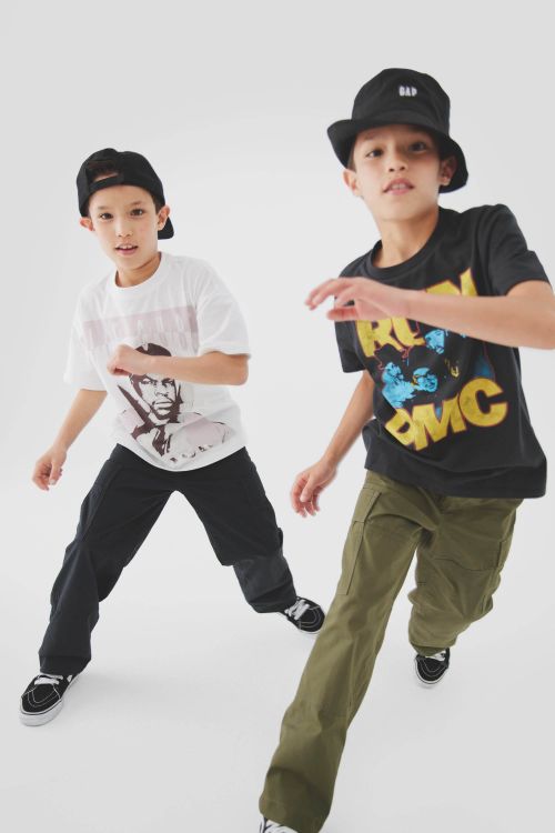 Two boys model youth fashion in a studio shot; one wears a Run DMC tee, bucket hat, and olive pants, while the other sports a portrait tee.