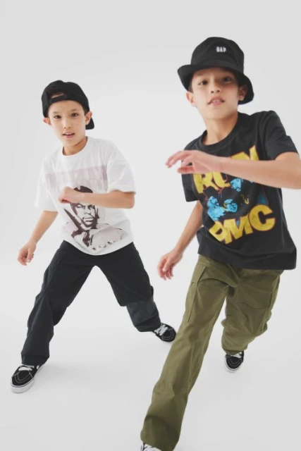 Two boys model youth fashion in a studio shot; one wears a Run DMC tee, bucket hat, and olive pants, while the other sports a portrait tee.