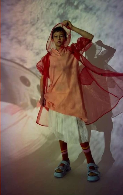 A young person poses in a hooded coral dress with a sheer red cape, white skirt, striped socks, and blue sandals against a soft, airy background.