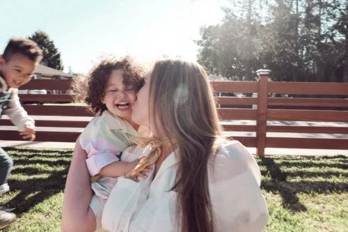 Laughing toddler with curly hair is kissed by an adult female in a sunny outdoor setting with a running child in the background.