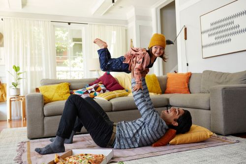 Father and daughter playing indoors, the dad lying on the floor holding his daughter in the air, with a pizza box nearby.