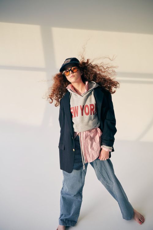 Stylish young person in a gray sweatshirt with 'New York' text, a blazer, and striped pants, standing in a naturally lit studio.