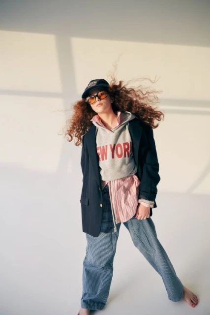 Stylish young person in a gray sweatshirt with 'New York' text, a blazer, and striped pants, standing in a naturally lit studio.