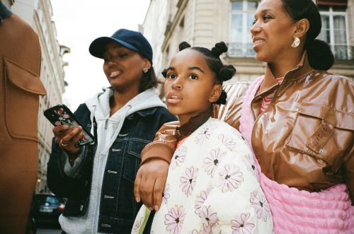 Three fashionable women of color walk down a European city street; a child with space buns wears a floral print jacket.