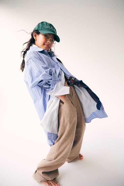 A young girl in an oversized blue shirt, khaki pants, and a green cap poses in a photo studio against a bright white background.