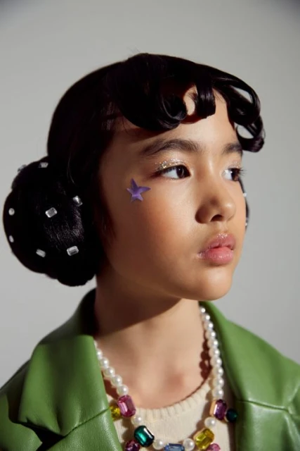 Young girl with glitter eyeshadow and a star on her cheek, accessorized with a pearl necklace and a green jacket, posing indoors.