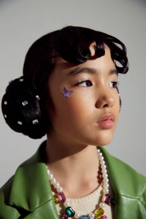 Young girl with glitter eyeshadow and a star on her cheek, accessorized with a pearl necklace and a green jacket, posing indoors.