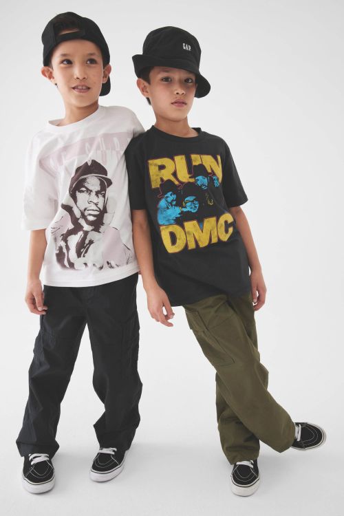 Two young boys pose in front of a white backdrop, wearing hats and graphic t-shirts featuring hip-hop artists and groups. 
