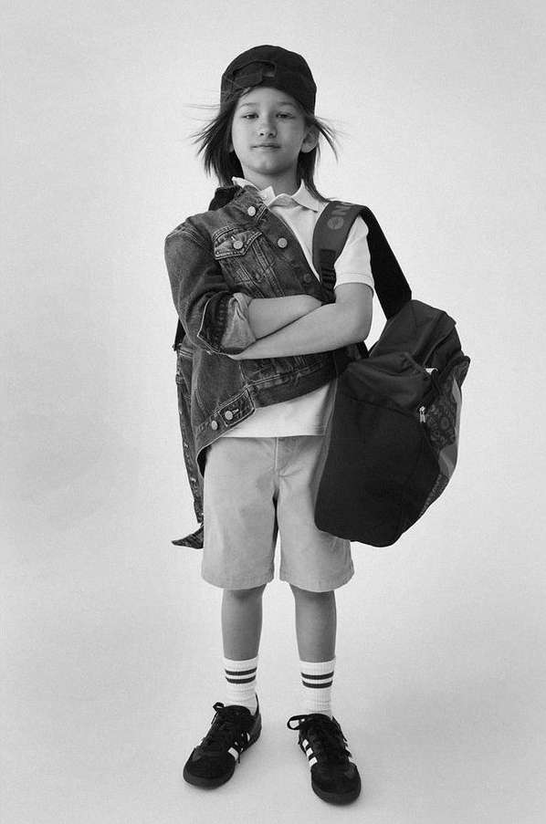 Child model wearing backwards cap, denim jacket, shorts, and carrying a backpack against a seamless backdrop