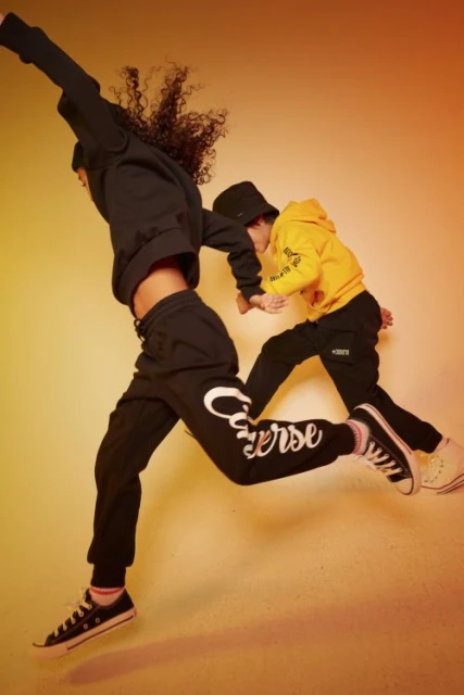Two kids in mid-air wearing Converse apparel: one in a black hoodie and pants, the other in a yellow hoodie and black pants.