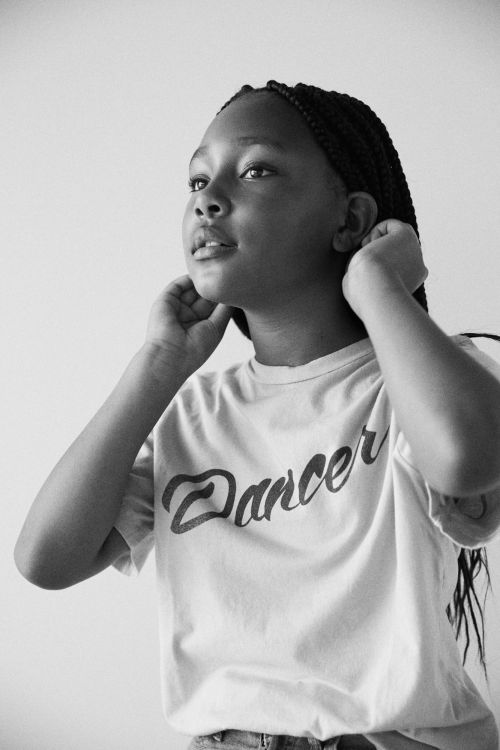 Black and white portrait of a young woman with braided hair wearing a 'Dancer' t-shirt, looking up with a gentle expression against a plain background.