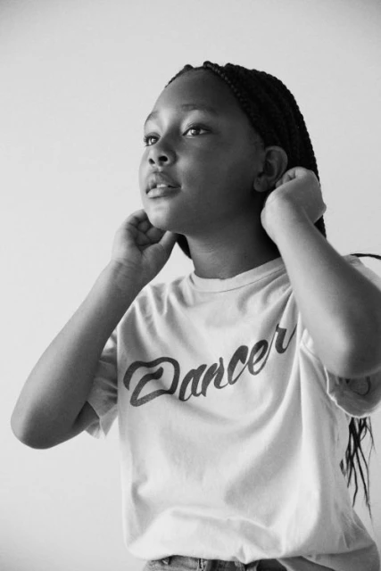 Black and white portrait of a young woman with braided hair wearing a 'Dancer' t-shirt, looking up with a gentle expression against a plain background.