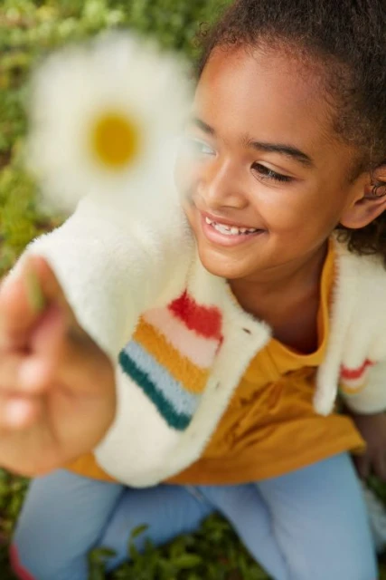 Smiling girl holds out white daisy in a sunny outdoor shot. Child wears colorful sweater and jeans, playing in green grass. 