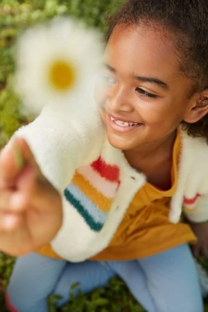 Smiling girl holds out white daisy in a sunny outdoor shot. Child wears colorful sweater and jeans, playing in green grass. 
