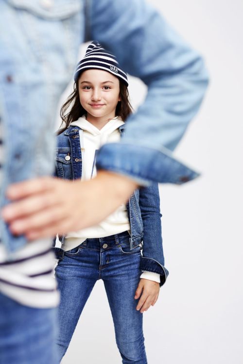 A young girl in a denim jacket and striped hat stands out against a clean backdrop, with another figure partially blurred in the foreground.