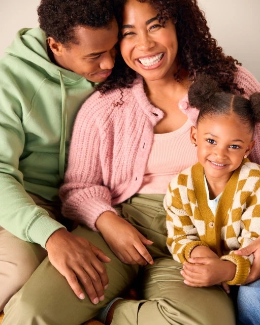 Smiling family portrait of a mother, father, and daughter indoors, wearing pastel colors and casual outfits in soft, diffused lighting.