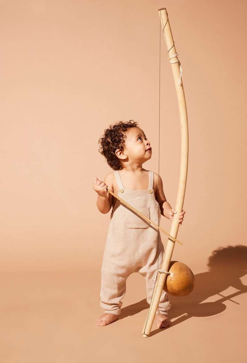 Toddler with dark curly hair stands holding a berimbau, a single-stringed percussion instrument from Brazil, looking up in studio.