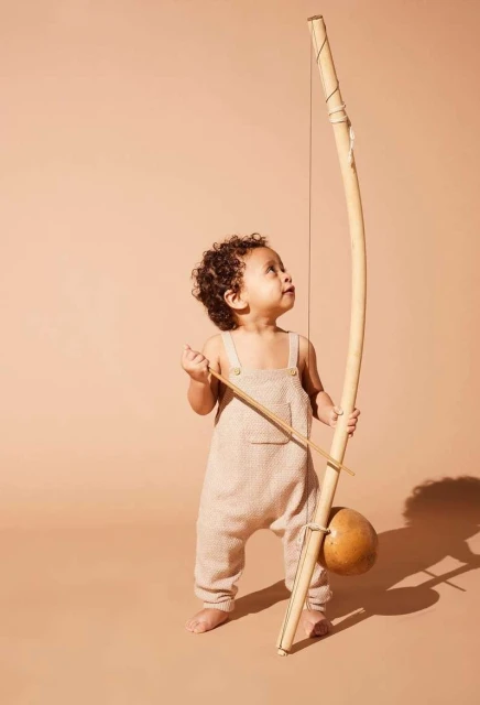 Toddler with dark curly hair stands holding a berimbau, a single-stringed percussion instrument from Brazil, looking up in studio.