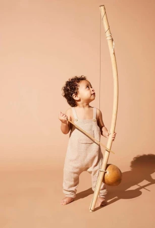 Toddler with dark curly hair stands holding a berimbau, a single-stringed percussion instrument from Brazil, looking up in studio.