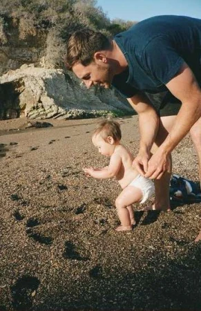 Father assists toddler taking first steps on a sandy beach near cliffs, bathed in soft sunlight, creating a heartwarming family moment.
