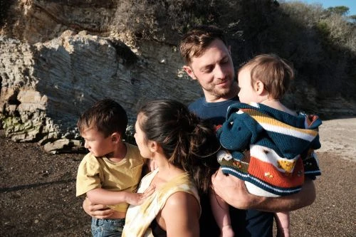 A family of four stands on a beach with rock formations in the background. The father holds a baby, while the mother and son stand beside them.