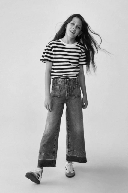 A young girl in a striped T-shirt and wide-leg jeans stands in a studio setting, casting a shadow on the white backdrop.
