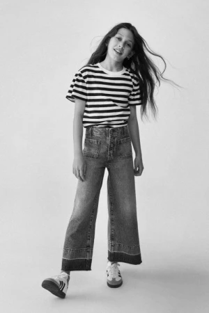 A young girl in a striped T-shirt and wide-leg jeans stands in a studio setting, casting a shadow on the white backdrop.