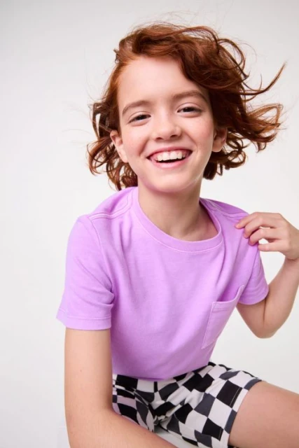 Cheerful child model in lilac t-shirt and checkered shorts smiles against a white background, showcasing vibrant youthful fashion.