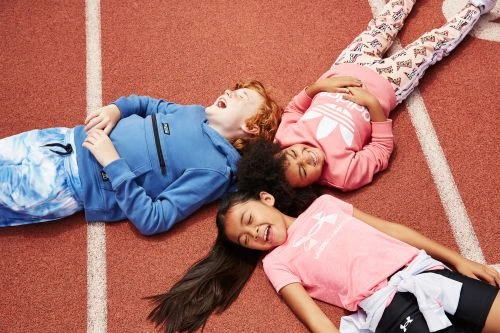 Three laughing children in athletic wear lie head-to-head on a red running track, wearing sweatshirts and leggings. Overhead shot, eye-level angle.