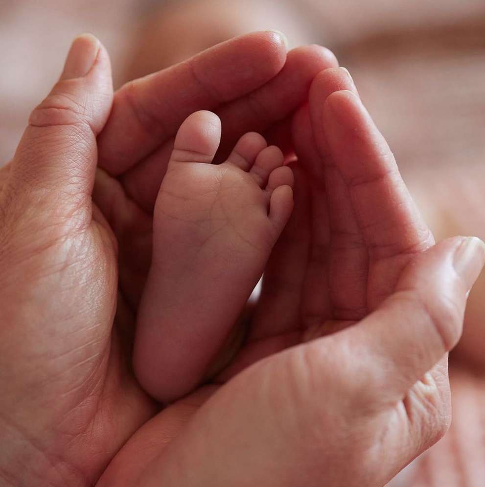 A baby's foot is cradled in adult hands, in a close-up shot with shallow depth of field and pink-toned hues, conveying tenderness.