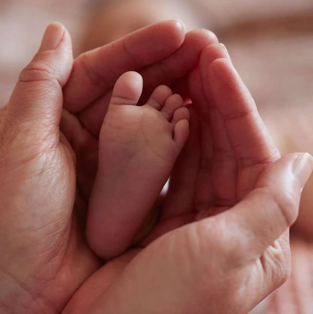 A baby's foot is cradled in adult hands, in a close-up shot with shallow depth of field and pink-toned hues, conveying tenderness.