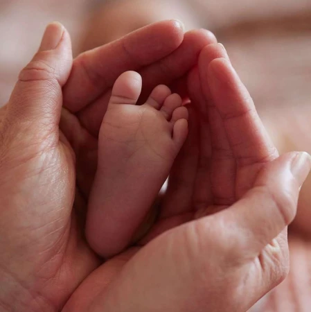 A baby's foot is cradled in adult hands, in a close-up shot with shallow depth of field and pink-toned hues, conveying tenderness.